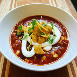 This image shows a bowl of taco soup topped with sour cream, cheese, corn chips, and cilantro microgreens.